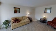 This single-floor space features a living room that serves as its central gathering area. The room is finished with wall-to-wall carpeting in a neutral beige tone, contributing to a warm and comfortable atmosphere. The walls are painted in a clean, crisp white, creating a bright and open feel throughout the space. A beige sofa anchors the seating area, accented with decorative cushions in deep red and burgundy tones, adding a subtle contrast to the neutral palette. A small dark wood side table positioned beside the sofa holds a classic table lamp, providing soft ambient lighting. On the opposite side of the room, a round dark wood coffee table completes the seating arrangement. Wall décor includes a framed piece of artwork depicting an urban cityscape in rich, warm tones, adding a distinctive visual focal point to the room. A standard interior door leads further into the space, suggesting additional rooms beyond the living area. The overall ambiance is modest and functional, with a clean, uncluttered layout that maximizes the usable floor area. The recessed ceiling vent indicates a central climate control system, ensuring year-round comfort.