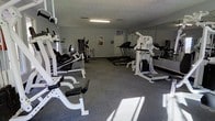 This single-floor fitness center is designed to support a full range of exercise routines in a functional and practical environment. The space is outfitted with an array of strength training equipment, including a multi-station cable machine, weight benches, and free-weight apparatus, providing users with comprehensive options for resistance training. On the cardio side, multiple treadmills are positioned along the room, offering ample availability during peak hours. A large wall mirror spans one side of the room, a classic feature that allows users to monitor their form and adds a sense of expanded space to the interior. The flooring consists of durable, cushioned gray carpet-style material well-suited for fitness use, offering both comfort underfoot and practical resilience. Overhead fluorescent lighting ensures the room is evenly and brightly illuminated throughout. The textured ceiling and neutral gray walls keep the aesthetic clean and utilitarian. Motivational posters and informational signage on the walls add a purposeful touch to the space. Overall, the fitness center provides a straightforward, no-frills environment focused entirely on delivering an effective and accessible workout facility.