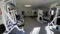 This single-floor fitness center is designed to support a full range of exercise routines in a functional and practical environment. The space is outfitted with an array of strength training equipment, including a multi-station cable machine, weight benches, and free-weight apparatus, providing users with comprehensive options for resistance training. On the cardio side, multiple treadmills are positioned along the room, offering ample availability during peak hours. A large wall mirror spans one side of the room, a classic feature that allows users to monitor their form and adds a sense of expanded space to the interior. The flooring consists of durable, cushioned gray carpet-style material well-suited for fitness use, offering both comfort underfoot and practical resilience. Overhead fluorescent lighting ensures the room is evenly and brightly illuminated throughout. The textured ceiling and neutral gray walls keep the aesthetic clean and utilitarian. Motivational posters and informational signage on the walls add a purposeful touch to the space. Overall, the fitness center provides a straightforward, no-frills environment focused entirely on delivering an effective and accessible workout facility.