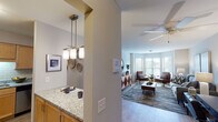 This thoughtfully designed single-floor residence offers a comfortable and functional layout across several well-defined living spaces. The open-concept kitchen is outfitted with warm maple cabinetry, granite countertops, a subway tile backsplash, stainless steel appliances including a dishwasher, and a double-basin sink — providing both style and practicality for everyday living. A peninsula with pendant lighting connects the kitchen to the dining area, creating a seamless flow between spaces.

The living room is a standout feature of the home, anchored by a large bay window arrangement that invites generous natural light and frames views of the surrounding greenery. Light hardwood-style flooring runs throughout this area, complementing the neutral wall tones and modern furnishings.

The residence includes two carpeted bedrooms, each offering a calm, restful atmosphere. The primary bedroom features ample closet space, while the secondary bedroom includes a built-in closet with light wood panel doors. Both rooms are served by their own full bathrooms, each finished with tile flooring, light wood vanities, and shower/tub combinations. A ceiling fan is installed in the living area, and the overall layout — as visible in the dollhouse view — is efficiently organized to maximize comfort and livability throughout.