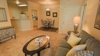 This single-floor residence offers a cohesive and inviting open-concept layout that seamlessly connects the living, dining, and kitchen areas into one harmonious living space. The living area is furnished with a plush, earth-toned sofa accented by patterned throw pillows and centered around an elegant oval glass-top coffee table, creating a comfortable and stylish gathering space. Warm, neutral carpeting flows throughout, contributing to the cozy ambiance of the interior.

Adjacent to the living area, the dining space features a classic round table surrounded by ornate wrought-iron chairs, set atop a dark area rug beneath a pendant light fixture that adds a refined focal point to the room. Framed artwork on the warm-toned walls enhances the traditional character of the space.

The kitchen is partially open to the main living area via a pass-through counter, allowing natural sightlines between rooms. It is fitted with light wood cabinetry, white appliances, and ample counter space, offering a functional and accessible cooking environment. Crown molding details along the ceilings add an architectural finishing touch throughout the space, while warm lighting creates an overall atmosphere that is both polished and livable.