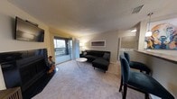 Located at 550 Harbor Cove Ln in Charleston, SC 29412, this single-floor residence offers a well-organized and functional layout designed for comfortable everyday living. The main living area is carpeted and spacious, centered around a sectional sofa and coffee table arrangement that makes efficient use of the open floor plan. A wall-mounted television and fireplace anchor one end of the room, while the opposite end opens to a private balcony through a sliding glass door, allowing natural light to fill the space and offering outdoor views of the surrounding harbor cove environment. Sheer curtains frame the balcony entrance, adding a soft, airy quality to the room. A textured ceiling runs throughout, and neutral wall tones keep the interior feeling bright and clean. A dedicated dining area is situated adjacent to the living room, furnished with teal accent chairs that introduce a pop of color. One of the standout features of this residence is the in-unit laundry area, visible from the living room and providing added convenience without leaving the home. The overall ambiance is casual and practical, with a layout that prioritizes open sight lines and ease of movement throughout the single-floor living space.