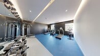 This single-floor fitness center offers a well-appointed workout environment thoughtfully designed within a compact footprint. The space is divided into two functional zones: a cardio area and a strength training area. The cardio zone features True-brand treadmills, elliptical trainers, and a stationary bike arranged along dark charcoal-painted walls, with natural light filtering in through windows that provide outdoor views. The strength training zone is anchored by a multi-function cable machine and a tiered dumbbell rack, complemented by a full-length mirror that visually expands the space. A standout design element is the dramatic three-dimensional geometric accent wall, featuring angular, faceted panels in a dark metallic finish that runs alongside the mirror, creating a bold, sculptural focal point. The ceiling incorporates recessed LED spotlights alongside continuous cove lighting at the perimeter, delivering layered, even illumination throughout. Blue rubber flooring transitions to light-toned tile in the weight area, clearly delineating each zone. The overall aesthetic is sleek and contemporary, combining functional fitness infrastructure with refined architectural detailing.