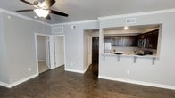 This single-floor space showcases an open-concept design that seamlessly connects the living area to the kitchen. Dark hardwood floors run throughout, adding warmth and contrast against the light grey walls and crisp white crown molding and baseboards. A ceiling fan with a light fixture anchors the living area, contributing to both comfort and style.

The kitchen is defined by rich, dark cabinetry that extends to the upper walls, paired with a light-toned granite or stone countertop that wraps around a peninsula-style breakfast bar — an ideal spot for casual dining or meal prep. A stainless steel refrigerator is visible, contributing a modern touch to the cooking space. Recessed lighting illuminates the kitchen efficiently.

A hallway visible from the main living area leads to additional rooms, with doors suggesting closet storage and separate bedroom access. The open flow between spaces creates a sense of generous square footage, while the consistent flooring and neutral color palette throughout lend the space a cohesive, well-maintained aesthetic. Overall, the layout prioritizes functionality while maintaining a clean, contemporary feel.