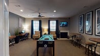 This single-floor amenity space is thoughtfully designed to serve as a multifunctional hub for recreation, relaxation, and productivity. The main gathering area centers around a full-size pool table set atop plush carpeted flooring, making it an inviting focal point for social activity. A wall-mounted flat-screen TV adds an entertainment element, while a fireplace along one wall lends warmth and character to the room. Seating is arranged throughout in the form of cushioned accent chairs and high-top bistro tables with industrial-style stools, providing versatile options for lounging or casual conversation. Natural light filters in through multiple windows dressed with navy blue curtains and horizontal blinds, complementing the neutral gray wall tones and creating a balanced, contemporary ambiance. A ceiling fan with integrated lighting keeps the space comfortable. A dedicated workstation nook features a wooden desk with a computer terminal, ideal for remote work or browsing. Adjacent to the main lounge, a glass-partitioned fitness center is visible and equipped with cardio machines, including treadmills, offering a convenient exercise option. Abstract framed artwork displayed in a triptych arrangement adds a polished, curated aesthetic throughout the space, tying the modern design together cohesively.