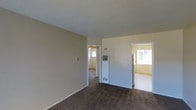 This single-floor residence offers a straightforward and functional layout with a comfortable, understated aesthetic. The main living area is finished with wall-to-wall carpet in a warm, neutral tone, complemented by smooth beige walls and white baseboards that lend a clean, cohesive look throughout. A wall-mounted climate control unit is centrally positioned, providing practical heating and ventilation for the space. The room connects openly to adjacent areas, with two doorways leading to additional rooms, each receiving generous natural light through windows — creating a bright and airy feel across the interior. The adjoining rooms continue the same carpeted flooring and neutral color palette, maintaining a consistent and unified design language throughout the home. The layout flows naturally from one room to the next, offering a connected and easy-to-navigate floor plan. Overall, the space presents a simple, livable environment with functional features and a neutral design well-suited for a variety of furnishing styles.