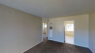 This single-floor residence offers a straightforward and functional layout with a comfortable, understated aesthetic. The main living area is finished with wall-to-wall carpet in a warm, neutral tone, complemented by smooth beige walls and white baseboards that lend a clean, cohesive look throughout. A wall-mounted climate control unit is centrally positioned, providing practical heating and ventilation for the space. The room connects openly to adjacent areas, with two doorways leading to additional rooms, each receiving generous natural light through windows — creating a bright and airy feel across the interior. The adjoining rooms continue the same carpeted flooring and neutral color palette, maintaining a consistent and unified design language throughout the home. The layout flows naturally from one room to the next, offering a connected and easy-to-navigate floor plan. Overall, the space presents a simple, livable environment with functional features and a neutral design well-suited for a variety of furnishing styles.