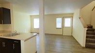 This two-floor space offers a clean, functional layout designed for comfortable everyday living. The main level features an open-concept design that seamlessly connects the kitchen and living areas, creating a spacious and versatile environment. The kitchen includes a peninsula counter that provides both workspace and a natural divider between the cooking and living zones, with light-toned countertops and a structural column adding architectural character to the space.

The living area is bright and welcoming, benefiting from multiple windows that allow generous natural light to fill the room. Warm, hardwood-style laminate flooring runs throughout the main level, adding a consistent and modern aesthetic. The entry door, trimmed in natural wood tones, opens directly into the living space and is flanked by a window, further enhancing the sense of openness and light.

The soft, neutral wall tones throughout complement the light flooring, creating a calm and versatile backdrop suitable for a variety of interior styles. The clean lines and unfussy design make the space highly adaptable. With a second floor adding additional living or sleeping quarters, this property offers a practical multi-level layout that balances open communal areas with the privacy of an upper level.