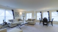 This two-floor residence offers a thoughtfully designed layout that balances comfort and elegance. The living area serves as the heart of the home, featuring crown molding, smooth ceilings, and neutral-toned walls that create a bright and airy atmosphere. Large windows dressed with layered curtains in dark and sheer fabrics allow abundant natural light to fill the room while adding a refined decorative touch. The carpeted living room accommodates generous seating arrangements, with ample space for a glass-top coffee table and accent pieces. Decorative wall art with ornate metalwork detailing adds character to the space. A dining area sits adjacent to the living room, furnished with dark wood high-back chairs, seamlessly integrating with the open-concept flow. The overall color palette of soft creams and warm neutrals lends a cohesive, sophisticated feel throughout the main level. The two-floor configuration provides a clear separation between living and private spaces, offering both functionality and privacy. With its classic architectural details, well-proportioned rooms, and carefully considered design elements, this residence presents a welcoming and polished environment suitable for both everyday living and entertaining.