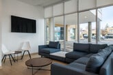 This contemporary single-floor communal space is defined by its clean, modern aesthetic and an abundance of natural light streaming through floor-to-ceiling glass curtain walls that frame views of the surrounding streetscape. Light-toned large-format tile flooring runs throughout, reinforcing the bright and open atmosphere.

The space is organized into several distinct functional zones. A casual lounge area features a generous sectional sofa and accent chairs arranged around a low coffee table, accompanied by a wall-mounted television — ideal for relaxed gatherings. Adjacent to this is a more intimate seating nook positioned directly against the expansive glazing, offering a round table and upholstered chairs bathed in natural light.

A vibrant social lounge sets itself apart with a bold red geometric-patterned area rug, mid-century-inspired armchairs with light wood frames, and a striking exposed brick accent wall that adds warmth and texture. A large pendant light anchors the seating arrangement, and glazed partitions provide a visual connection to neighboring areas.

The kitchenette is a standout feature, showcasing a dramatic terracotta-glazed tile backsplash, dark wood cabinetry, white quartz countertops, and a stainless steel refrigerator. A bar-height island with seating and suspended red pendant lights completes the space, offering a stylish spot for informal dining or conversation.