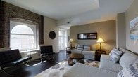 Located at 699 N Broad St in Philadelphia, PA, this single-floor space masterfully combines historic character with contemporary style. The living area immediately draws attention with its soaring ceilings and striking exposed brick column, a nod to the building's storied past. Large arched windows flood the space with abundant natural light, highlighting the rich dark hardwood floors that run throughout. The living area is thoughtfully arranged with a neutral-toned sectional sofa, a sleek round coffee table, and a classic Barcelona-style accent chair, all anchored by a patterned area rug that adds warmth and texture. A decorative wall mirror reflects the open layout, creating a sense of added depth, while warm table lamps contribute to a refined, welcoming ambiance. The open sightline to the kitchen, visible through a framed wall opening, suggests a functional and connected layout. Neutral wall tones throughout complement both the historic and modern elements, resulting in a cohesive aesthetic. The combination of arched architectural details, exposed masonry, and curated contemporary furnishings gives this Philadelphia space a distinctive character that balances urban sophistication with timeless design.