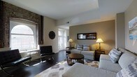 Located at 699 N Broad St in Philadelphia, PA, this single-floor space masterfully combines historic character with contemporary style. The living area immediately draws attention with its soaring ceilings and striking exposed brick column, a nod to the building's storied past. Large arched windows flood the space with abundant natural light, highlighting the rich dark hardwood floors that run throughout. The living area is thoughtfully arranged with a neutral-toned sectional sofa, a sleek round coffee table, and a classic Barcelona-style accent chair, all anchored by a patterned area rug that adds warmth and texture. A decorative wall mirror reflects the open layout, creating a sense of added depth, while warm table lamps contribute to a refined, welcoming ambiance. The open sightline to the kitchen, visible through a framed wall opening, suggests a functional and connected layout. Neutral wall tones throughout complement both the historic and modern elements, resulting in a cohesive aesthetic. The combination of arched architectural details, exposed masonry, and curated contemporary furnishings gives this Philadelphia space a distinctive character that balances urban sophistication with timeless design.