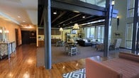 This single-floor space showcases a striking industrial loft aesthetic, defined by exposed dark steel beams and trusses that span the ceiling, creating a bold architectural statement. The open-concept layout flows effortlessly from a well-appointed kitchen area into a casual dining zone and a comfortable lounge space, making it ideal for both gathering and relaxing. Warm hardwood flooring anchors the entry area, transitioning into carpeted zones that delineate the dining and seating areas. The kitchen is outfitted with dark wood cabinetry and ample counter space, while pendant lighting with angular lamp shades adds a warm, inviting glow throughout. The dining area features round tables with high chairs, encouraging informal interaction, while the lounge is furnished with plush sofas and accent chairs arranged around a central coffee table. Large windows along the far wall allow natural light to pour in, offering views of the outdoors and brightening the interior. Exposed ductwork and structural columns reinforce the industrial character of the space, while warm yellow-toned walls soften the overall atmosphere. This thoughtfully designed communal space balances rugged architectural detail with comfortable, functional furnishings.