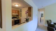 This two-story residence offers a functional and welcoming layout designed for comfortable everyday living. The kitchen is a central feature, equipped with warm-toned wood cabinetry, a black electric range with an overhead ventilation hood, and dark countertops. A built-in pass-through counter connects the kitchen to the adjacent hallway area, creating an open flow between spaces and allowing for easy interaction between rooms. Upper cabinets, including a peninsula-style overhead cabinet accessible from both sides of the pass-through, provide ample storage. The kitchen also includes a closet with louvered doors, offering additional organizational space. A hallway extends from the kitchen area, finished with neutral carpet flooring and light-toned walls, leading toward additional rooms visible in the background. The overall interior palette is kept light and neutral throughout, with flush-mount ceiling lighting providing consistent illumination. The layout emphasizes practicality and connectivity between living spaces, with the pass-through counter serving as both a visual and functional link across the main level of the home.