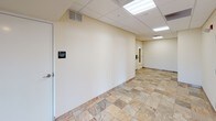 This single-floor commercial lobby and hallway space offers a clean, functional layout designed to serve as a welcoming entry point and circulation corridor. The flooring throughout is finished in large-format natural stone-look tiles in warm, variegated earth tones, lending the space a polished and durable character.

The corridor runs a generous length, with cream-painted walls accented by a subtle tile baseboard detail that ties the design together cohesively. A drop ceiling with a standard grid system is equipped with flush recessed fluorescent panels, providing consistent and even illumination across the full length of the space.

At one end, a glass-panel entry door allows natural light to filter into the corridor, creating a bright and inviting front entrance. Visible signage directs occupants to an elevator equipment room, indicating that the building supports vertical access infrastructure. A wall-mounted intercom and keypad entry system is positioned near the entrance, providing controlled access for building security.

A built-in mailbox unit with individual compartments is mounted along one wall, serving the building's residents or tenants with a convenient mail distribution point. French-style double doors along the corridor lead to an exterior area currently under construction, visible through safety fencing. The overall ambiance is practical and well-maintained, suited to a multi-tenant residential or mixed-use commercial building.