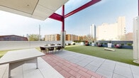 Located at 1717 N 12th St in Philadelphia's vibrant North Philadelphia neighborhood, this outdoor amenity terrace is a thoughtfully designed communal space offering a wide range of leisure and social features. The terrace is defined by its bold architectural aesthetic, with striking red structural columns and beams contrasting against clean white paneling and modern metallic finishes throughout.

A generous expanse of artificial turf forms the centerpiece of the terrace, providing a versatile open-air lawn ideal for recreation and relaxation. Dedicated zones for lawn games, including cornhole, are integrated into the space, while a ping-pong table occupies a covered patio section. An oversized wall-mounted outdoor television, positioned beneath a covered overhang with roll-down shutters, creates an entertainment hub suited for group gatherings.

Multiple seating arrangements are distributed across the terrace, including large L-shaped wicker sectionals with bold red cushions, bistro-height tables with red barstools, and intimate lounge chairs. Raised planters filled with decorative shrubs and greenery line the perimeter, softening the urban setting. The terrace offers open, panoramic views of the surrounding Philadelphia skyline and cityscape, flooding the space with natural light throughout the day.