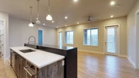 This single-floor space offers a thoughtfully designed open-concept layout that seamlessly connects the kitchen and living areas. The kitchen is a standout feature, showcasing a large island with crisp white quartz countertops and a richly textured dark wood base, complemented by a professional-style spring-neck faucet. Pendant lights with polished chrome fixtures hang above the island, adding both function and a modern industrial flair. Recessed lighting throughout the space ensures consistent, even illumination. The living area is bright and airy, with multiple windows dressed in horizontal blinds that allow generous natural light to fill the room. A ceiling fan overhead provides comfort and airflow. Light-toned hardwood flooring runs throughout, contributing warmth and continuity across the open floor plan. A glass-paneled exterior door provides additional light and access to the outdoors. The neutral color palette of soft beige walls and white trim creates a clean, welcoming ambiance that is both versatile and inviting. The overall design balances contemporary finishes with practical functionality, making for a comfortable and visually cohesive living environment.