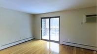 This single-floor space offers a clean, functional layout with notable architectural details. The main living area features polished hardwood flooring that reflects natural light, creating a warm and inviting atmosphere. Neutral-toned walls provide a versatile backdrop suitable for a variety of interior styles.

A prominent sliding glass door serves as a central architectural feature, opening onto an outdoor balcony enclosed by a metal railing. The balcony is partially framed by a decorative lattice panel on one side, adding a subtle design element. Through the glass door, views of surrounding trees and the outdoors are visible, enhancing the sense of connection to the exterior environment.

Baseboard heaters are positioned along multiple walls, ensuring efficient climate control throughout the space. The ceiling is smooth and finished in white, complementing the bright, airy feel of the room. The overall ambiance is understated and practical, with an emphasis on natural light and straightforward design. The space presents itself as a well-maintained, move-in-ready environment with solid foundational features.
