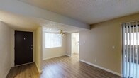 This single-floor space features an open and functional layout that makes efficient use of its footprint. The main living area is finished with smooth, wood-style laminate flooring that adds warmth and continuity throughout the space. A textured ceiling with a ceiling fan sits above, providing both visual character and practical air circulation. Natural light flows in through a large window dressed with vertical blinds, brightening the living space and creating an inviting atmosphere. Neutral-toned walls in a soft beige palette provide a clean, versatile backdrop throughout. A structural beam detail adds a subtle architectural element, subtly defining the transition between spaces. An open doorway connects the living area directly to the kitchen, which is visible in the background and features white cabinetry and what appears to be light countertops — keeping the overall aesthetic bright and cohesive. A dark-paneled entry door is visible at the side, marking the main entrance to the unit. The straightforward, open-plan design promotes easy flow between the living and kitchen areas, making the space well-suited for everyday living.
