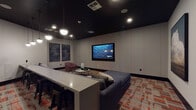 Located at 12500 SW 175th Ave in Beaverton, Oregon, this single-floor media lounge is designed with a sleek, contemporary aesthetic that prioritizes comfort and entertainment. The room is defined by its striking dark ceiling, which creates a dramatic contrast against the light grey walls and lends the space a cinematic, immersive atmosphere.

At the focal point of the room is a large wall-mounted flat-screen television, framed by vertically paneled wall treatments that add texture and visual depth. The seating arrangement includes a generously sized navy blue sectional sofa accented with grey and blush pink throw pillows, positioned to face the screen for optimal viewing. A burgundy accent chair and a round coffee table complete the lounge area.

A raised counter with bar-height seating separates a secondary zone from the main viewing area, offering a versatile space for casual use. Pendant globe lights hang from the ceiling, complementing the recessed lighting to provide layered illumination throughout. A bold, patterned carpet in shades of red, orange, and grey grounds the room with color and warmth.

Curated artwork adorns the walls, including a large-scale cloudscape painting and abstract blue panels, adding an artistic dimension to the space. A black barn-style sliding door provides a stylish entryway detail, reinforcing the room's modern design sensibility throughout.
