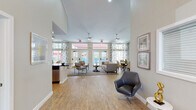 This single-floor communal space is defined by its striking architectural character and open, welcoming layout. The entryway sets an impressive tone immediately, featuring soaring vaulted ceilings, a elegant chandelier, recessed lighting, and a grand arched window above French doors that frame outdoor views and draw in generous natural light. Light wood-tone flooring runs throughout, complementing the neutral, soft white wall palette.

The main gathering area is expansive and thoughtfully arranged, anchored by a tufted sofa, accent chairs, and a decorative coffee table atop a patterned area rug. Dual ceiling fans overhead provide both visual interest and practical comfort. Floor-to-ceiling French doors with transom windows line the far wall, offering direct views and access to an outdoor pool area while flooding the interior with natural light. Floral-patterned curtains add a refined, residential touch alongside a curated gallery wall.

A dedicated dining area nearby features a round dark wood table surrounded by upholstered chairs, positioned beside large windows dressed with floral drapery. The kitchen is well-appointed with gray cabinetry, dark granite countertops, stainless steel appliances including a side-by-side refrigerator, range, and microwave, and light wood flooring continuing seamlessly throughout. The overall ambiance is polished, bright, and inviting.