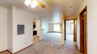 This single-floor space features an open layout that connects the main living area to the kitchen and additional rooms. The living area is finished with wall-to-wall carpet and topped by a textured ceiling, with a ceiling fan providing both lighting and air circulation. A sliding glass door and a side window allow natural light to fill the space, creating a bright and airy atmosphere. A baseboard heating unit runs along the exterior wall beneath the window, ensuring comfortable temperatures year-round. The kitchen, partially visible through an open doorway, includes dark wood cabinetry, a black electric range, and a built-in microwave above the stove, with resilient flooring that contrasts the carpeted living areas. An electrical panel is mounted on the wall near the kitchen entry, offering convenient access. A structural column adds architectural definition to the open floor plan. A hallway visible on the opposite side leads to additional rooms, with wood-toned doors and trim providing warmth throughout. The overall design is functional and straightforward, with a neutral color palette that makes the space feel clean and versatile.