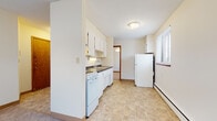 This single-floor unit offers a straightforward, functional layout with clearly defined living, kitchen, and bathroom areas. The kitchen is equipped with white cabinetry extending to the ceiling, providing ample storage space. A coil-burner electric range, a double-basin stainless steel sink with an under-cabinet range hood, dark countertops, and a full-size refrigerator complete the kitchen's practical setup. The flooring in the kitchen features neutral tile in a diagonal pattern, contributing to a clean, utilitarian aesthetic.

The living area and bedroom spaces are finished with wall-to-wall carpet in a neutral beige tone, lending a modest warmth to each room. Windows fitted with horizontal blinds allow natural light to filter in, with views of the surrounding exterior. Baseboard heating units run along the lower walls of the carpeted rooms, providing climate control throughout the unit.

The bathroom includes a pedestal sink, a medicine cabinet with mirror, and a full soaking tub surrounded by white ceramic tile. The overall palette of the unit is light and neutral, with cream-colored walls throughout, creating a simple, move-in-ready environment suited for everyday living.