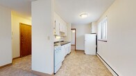 This single-floor unit offers a straightforward, functional layout with clearly defined living, kitchen, and bathroom areas. The kitchen is equipped with white cabinetry extending to the ceiling, providing ample storage space. A coil-burner electric range, a double-basin stainless steel sink with an under-cabinet range hood, dark countertops, and a full-size refrigerator complete the kitchen's practical setup. The flooring in the kitchen features neutral tile in a diagonal pattern, contributing to a clean, utilitarian aesthetic.

The living area and bedroom spaces are finished with wall-to-wall carpet in a neutral beige tone, lending a modest warmth to each room. Windows fitted with horizontal blinds allow natural light to filter in, with views of the surrounding exterior. Baseboard heating units run along the lower walls of the carpeted rooms, providing climate control throughout the unit.

The bathroom includes a pedestal sink, a medicine cabinet with mirror, and a full soaking tub surrounded by white ceramic tile. The overall palette of the unit is light and neutral, with cream-colored walls throughout, creating a simple, move-in-ready environment suited for everyday living.