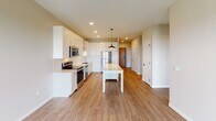 Located at 1408 Second St SW in Rochester, Minnesota, this thoughtfully designed single-floor residence combines modern finishes with a functional, open layout. The heart of the unit is a spacious open-concept kitchen, showcasing crisp white shaker cabinetry that reaches the ceiling, a classic white subway tile backsplash, light quartz countertops, and a full suite of stainless steel appliances including a range, microwave, dishwasher, and refrigerator. A central island with built-in wine storage and a stylish pendant light above serves as both a prep space and a casual gathering point. Warm-toned luxury vinyl plank flooring flows throughout the main living areas, creating a cohesive and inviting atmosphere. A wide hallway connects the living spaces to the private rooms, lined with clean white panel doors and black hardware accents. The bedroom is generously sized with plush carpeting and a large picture window framing views of the surrounding neighborhood, allowing abundant natural light to fill the room. The bathroom is finished with a white vanity, a framed mirror, and a full tub-shower combination. A dedicated laundry room houses a stacked Whirlpool washer and dryer alongside built-in wire shelving for additional storage. The overall aesthetic is clean, contemporary, and move-in ready.