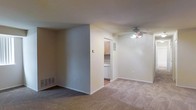 This single-floor residence offers a straightforward and functional layout designed for comfortable everyday living. The main living area is finished with wall-to-wall carpeting in a neutral tone, creating a warm and cohesive feel throughout the space. Smooth, light-colored walls provide a clean backdrop, and a ceiling fan with integrated lighting is centrally positioned overhead, ensuring both air circulation and ambient illumination. A floor vent indicates a central HVAC system for year-round climate control. A partial wall divides the living area from the kitchen, offering a degree of separation while maintaining an open connection between the two spaces. The kitchen features light wood cabinetry and white appliances, contributing to a bright and tidy aesthetic. The layout is efficient, making good use of the available footprint. White baseboards trim the perimeter of the rooms, adding a crisp finishing detail throughout. Overall, the space presents a practical and move-in-ready environment with a neutral palette that lends itself well to a variety of interior styles and personal touches.