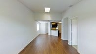 This single-floor residence is designed with an open, airy layout that seamlessly connects the main living area to the kitchen. The living space is finished with warm-toned hardwood flooring that extends throughout, creating a cohesive and inviting atmosphere. A centrally positioned skylight floods the room with natural light, complementing the clean white walls and bright ceiling to enhance the sense of openness.

The kitchen is partially open to the living area, separated by a half-wall peninsula that features a granite-style countertop — ideal as a breakfast bar or additional prep surface. Dark espresso-toned cabinetry provides a striking contrast against the lighter surroundings, adding depth and a modern aesthetic. A white refrigerator is visible within the kitchen, and overhead lighting keeps the space well-illuminated.

A window positioned along the living area wall introduces additional natural light, while multiple doorways lead to other areas of the home, suggesting further rooms beyond the main living space. The overall design is clean and functional, with a neutral color palette that offers versatility for a variety of interior styles. The layout and finishes combine to create a practical yet stylish single-floor living environment.
