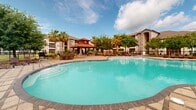 Located at Belmere Luxury Court in Houma, Louisiana, this outdoor amenity space centers around a generously sized freeform swimming pool with a striking turquoise water finish. The pool's organic, curvilinear shape creates a resort-like atmosphere, complemented by a dedicated wading area or spa feature visible at one end. The surrounding deck is finished in a durable, earth-toned paver design that extends broadly in all directions, providing ample room for lounging and socializing. Numerous poolside chaise lounges and seating arrangements are positioned throughout the deck, offering residents plenty of space to relax. A covered gazebo with a distinctive red metal roof serves as a shaded gathering spot and architectural focal point within the courtyard. Mature palm trees and other lush greenery are interspersed around the perimeter, lending a tropical ambiance and natural privacy. The space is enclosed by a decorative iron fence, ensuring both safety and a defined boundary. The multi-story residential buildings surrounding the pool area feature a mix of brick and neutral siding exteriors with pitched rooflines, providing a pleasant architectural backdrop that enhances the overall sense of a well-appointed, amenity-rich community.