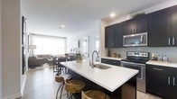 This single-floor space is designed around an open-concept layout that integrates the kitchen, dining, and living areas into one cohesive, functional environment. The kitchen serves as the focal point, showcasing dark, flat-front cabinetry that contrasts sharply with the bright white quartz countertops. A generously sized island anchors the kitchen, equipped with an undermount sink, a gooseneck faucet, and seating for multiple guests via rustic wooden barstools that add a warm, organic contrast to the modern aesthetic. Stainless steel appliances, including a range, microwave, and additional cooking appliances, line the back wall alongside a light grey subway tile backsplash that ties the design together. The open layout transitions naturally into the dining area, where a dining table and chairs are positioned to take advantage of natural light streaming in through large windows. Beyond, the living area provides a comfortable and relaxed retreat. Recessed lighting throughout ensures consistent illumination, while the light hardwood flooring and neutral walls create a bright, airy ambiance across the entire space.