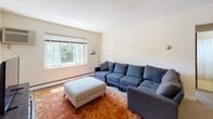 Located at 12 Brattle Dr in Arlington, MA, this single-floor residence offers a well-organized layout designed for comfortable everyday living. The living area is generously proportioned, featuring soft carpeting, neutral cream-toned walls, and a large window that draws in natural light and offers views of surrounding greenery. A open shelving divider subtly separates the living space from a dedicated dining area, providing definition without sacrificing openness. The kitchen is thoughtfully equipped with warm oak cabinetry, tile backsplash, white countertops, a stainless steel sink, and white appliances including a dishwasher. A window above the counter fills the kitchen with natural light, while tile flooring adds durability and ease of maintenance. The bedrooms are carpeted and calm, each finished with cream walls and wood-toned doors. One bedroom benefits from dual windows that bring in ample light and views of the exterior brick facade. The bathroom features classic mosaic tile flooring, a shower-tub combination, and a clean, functional layout. Baseboard heating is present throughout, ensuring year-round comfort. Overall, the space presents a practical and livable floor plan with a warm, understated aesthetic.