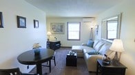 This single-floor space features an open-concept layout that combines a living area and a dining nook into one cohesive, functional room. The living area is anchored by a light-toned sectional sofa accented with teal and striped throw pillows, paired with a dark upholstered ottoman that doubles as a coffee table. A wall-mounted mini-split air conditioning unit ensures year-round climate comfort. Natural light enters through a double-hung window with dark wood trim, brightening the neutral, light-colored walls. The carpeted flooring runs throughout, adding warmth underfoot. Framed artwork adorns the walls, contributing a decorative, curated feel to the space. The dining area features a dark, round bistro-style table with matching chairs, ideal for intimate meals. Additional furnishings include a dark media console and coordinating side tables. A floor lamp and torchiere lamp provide layered ambient lighting options. The textured ceiling adds a subtle architectural detail. Overall, the space presents a compact yet well-organized layout with a clean, understated aesthetic suited for comfortable everyday living.