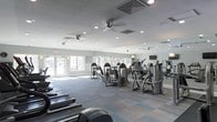 This single-floor fitness center is designed to accommodate a comprehensive workout experience, offering a well-rounded selection of both strength training and cardiovascular equipment. The space features a variety of selectorized strength machines arranged throughout the room, alongside cardio equipment including treadmills, ellipticals, and additional aerobic machines positioned to maximize the available floor space.

The interior is finished with a patterned, multi-tone blue and gray carpet that adds a functional yet contemporary aesthetic to the space. Walls are painted in a neutral light gray, contributing to a clean and modern atmosphere. Full-length mirrors line one side of the room, visually expanding the space and allowing users to monitor their form during workouts.

Overhead, recessed lighting combined with ceiling fans ensures the space remains well-lit and comfortable throughout use. Natural light filters in through windows along the exterior wall, enhancing the overall brightness of the room. A mounted flat-screen television is also visible, providing entertainment options for those using the cardio equipment. The layout is open and spacious, allowing for easy movement between stations.