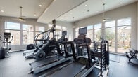 Fitness Center 2 is a thoughtfully designed, single-floor facility that offers a comprehensive workout environment for residents. The space is divided into two distinct zones: a cardio area and a strength-training area, each fully equipped with professional-grade machines.

The cardio section features multiple Cybex treadmills and StairMaster units, each equipped with individual digital display screens. This area is lined with large, multi-pane windows that flood the space with natural light and offer views of the surrounding community, including greenery and a pool area. Recessed ceiling lights and pendant fixtures provide additional illumination, while a ceiling fan ensures comfortable airflow.

The strength-training area is equally well-appointed, housing a range of cable machines, weight benches, a dumbbell rack, and multi-station resistance equipment from brands including Magnum. High ceilings amplify the sense of openness in this section, and expansive floor-to-ceiling windows continue the theme of natural light and outdoor connectivity.

Throughout the facility, durable rubber flooring provides safety and comfort underfoot. The clean, neutral color palette of warm gray walls and modern finishes creates a focused, professional atmosphere. The overall layout is spacious and organized, making it easy to navigate between equipment and supporting an effective, uninterrupted workout experience.
