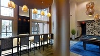 This sophisticated single-floor space combines modern design with refined architectural details to create a welcoming and visually striking environment. At its centerpiece is a dark granite-topped bar counter lined with cushioned high-back stools, ideal for casual seating or social gatherings. Above, a striking cluster of sculptural gold pendant lights casts a warm, ambient glow that enhances the space's upscale character. A prominent textured cylindrical column anchors the layout, adding architectural interest and visual depth. Floor-to-ceiling windows along the perimeter flood the interior with natural light while framing views of the surrounding brick exterior, creating a seamless connection between indoors and out. Sheer drapery softens the window line, adding an elegant touch to the bright, airy atmosphere. Toward the rear of the space, a cozy seating area sits atop a deep blue area rug, complemented by tall decorative greenery in a sleek white planter. A fireplace feature along the far wall adds warmth and a refined focal point. The overall ambiance is polished and inviting, blending contemporary furnishings with thoughtful lighting and architectural elements to deliver a comfortable yet stylish communal environment.