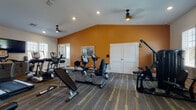 Located at 726 Cumberland Trace Rd in Bowling Green, Kentucky, this well-appointed fitness center is designed to accommodate a wide range of workout routines in a comfortable, single-floor layout. The main exercise area features striking vaulted ceilings accented by a bold amber-orange feature wall, ceiling fans, and recessed lighting that create an energizing yet welcoming atmosphere. The space is divided into distinct functional zones, with durable striped carpet flooring in the equipment areas and rich hardwood-style flooring in the cardio section. The cardio zone is equipped with multiple TRUE-brand treadmills and stair climbers, all positioned to face large windows fitted with white blinds that invite ample natural light and outdoor views. A wall-mounted flat-screen TV enhances the workout experience. The strength and cycling area houses upright and recumbent stationary bikes, a weight bench, and a cable resistance machine, offering comprehensive training options. A separate studio room provides a quieter space for flexible workouts, complete with an elliptical, exercise mat, stability ball, and a wall-mounted display screen. Full-length mirrors in the main area allow users to monitor their form. The facility connects to an adjacent corridor, suggesting integration within a larger community amenity complex.