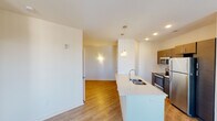 This single-floor residence offers a thoughtfully designed layout that balances modern finishes with functional living spaces. The kitchen is a standout feature, showcasing stainless steel appliances including a refrigerator, range, and microwave, complemented by sleek dark cabinetry with contemporary hardware and light quartz countertops. A peninsula with an integrated sink and pendant lighting above adds both style and utility, creating a natural connection to the open living and dining areas. Light wood-tone flooring runs throughout the main living spaces, lending warmth and continuity to the open floor plan.

The living area benefits from multiple windows fitted with horizontal blinds, allowing ample natural light to fill the space, along with a glass exterior door that provides additional brightness and outdoor access. The bedroom is finished with plush carpeting and features a large window with views to the surrounding neighborhood. An adjacent walk-in closet offers generous storage with wire shelving, while the bathroom includes a soaking tub with shower, tile surround, and built-in shelving niches. A secondary bathroom presents a clean, minimal design with a round mirror, pedestal sink, and gold-toned hardware accents. Throughout, the interiors are finished in crisp white tones, creating a bright and inviting atmosphere.