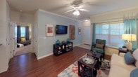 This single-story residence is thoughtfully designed with a functional layout and warm, neutral finishes throughout. The living room serves as a welcoming central space, featuring rich hardwood-style flooring, crown molding, a ceiling fan, and soft blue accent walls. Natural light filters through large windows dressed with grommet-panel curtains, creating a bright and comfortable atmosphere.

The galley-style kitchen is efficiently arranged with warm honey-toned cabinetry, dark granite countertops, light tile flooring, and black appliances including a range, microwave, refrigerator, and dishwasher. Ample upper and lower cabinet storage ensures generous prep and storage space.

The primary bedroom is generously sized, showcasing plush carpet, a vaulted ceiling with a ceiling fan, warm golden accent walls, and a large window allowing natural light. It connects to a spacious walk-in closet outfitted with extensive wire shelving systems and hanging rods. A second carpeted bedroom offers similar comfort with ceiling fan and closet access.

The bathroom features tile flooring, a tub-shower combination, a dark granite vanity top, and wood cabinetry, maintaining the consistent, polished aesthetic found throughout the residence.