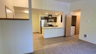This single-floor space showcases a thoughtfully designed open-concept layout that seamlessly connects the living and kitchen areas. The kitchen is a standout feature, equipped with stainless steel appliances including a refrigerator, microwave, and range. Dark espresso-toned upper cabinetry contrasts cleanly against light countertops and white lower cabinetry, creating a modern two-tone aesthetic. A peninsula with a white waterfall-style surround provides additional prep space and serves as a natural divider between the kitchen and living area. Recessed lighting throughout the kitchen ensures the space is well-lit. The flooring transitions from light wood-look planks in the kitchen and main area to carpet in the living zone, adding warmth underfoot. Structural columns frame the open pass-through, giving the layout a defined yet airy feel. A dedicated in-unit laundry area is visible to the side, offering added convenience with a full-size washer and dryer. The overall palette of warm neutrals and clean finishes gives the space a bright, contemporary ambiance suited for comfortable everyday living.
