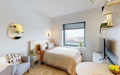 This thoughtfully designed single-floor studio maximizes every square foot through smart spatial planning and a cohesive modern aesthetic. Light wood flooring runs throughout the main living area, complementing the crisp white walls and creating a bright, airy atmosphere.

The sleeping area is anchored by a large picture window fitted with a roller shade, offering elevated views of the surrounding cityscape. A wall-mounted sconce provides warm accent lighting, while a compact armchair adds a functional seating nook. A decorative round mirror enhances the sense of openness in the room.

A full-height built-in wardrobe system with integrated open shelving serves as both a storage solution and a subtle room divider between the sleeping area and the bathroom. The shelving nooks offer accessible display and storage space.

The kitchenette is streamlined and efficient, featuring flat-front white cabinetry extending from floor to ceiling, an induction cooktop, an undermount sink, and a built-in microwave. Under-cabinet lighting illuminates the clean white countertop workspace.

The bathroom is finished in soft sage-green tones with light blue square floor tiles. It includes a wall-mounted sink, a wall-hung toilet, a mirrored medicine cabinet, and a heated towel rail, creating a calm, spa-like atmosphere within a compact footprint.