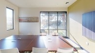 This student lounge is a well-appointed, single-floor space designed to support collaborative study and group gatherings. At the center of the room sits a large rectangular wood-toned conference table surrounded by white chairs, providing ample seating for group work or meetings. The floor is covered in a bold, multicolored patterned carpet featuring shades of blue, yellow, and gray, adding a lively visual element to the space.

Natural light flows generously through large floor-to-ceiling windows along one wall, offering views of the surrounding outdoor landscape, including mature trees and an exterior courtyard area. The bright, open feel is complemented by recessed ceiling lighting and clean white walls.

A wall-mounted flat-screen television is positioned prominently on one wall, making the lounge suitable for presentations or group viewings. Decorative horizontal artwork with a rich, multicolored palette adorns the walls, enhancing the creative and welcoming atmosphere. An acoustic panel on one wall contributes to sound management within the space.

The lounge is accessible via a doorway that connects to a corridor beyond, with an illuminated exit sign indicating clear egress. Overall, the space strikes a balance between functional design and an inviting, student-friendly aesthetic.