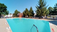 Located at 3675 Cross Ridge Ln in Canal Winchester, Ohio, this single-level community amenity space is centered around a generously sized outdoor swimming pool with crystal-clear turquoise water. The pool area is thoughtfully designed for relaxation and recreation, featuring a classic metal entry rail, poolside lounge chairs, and a smooth surrounding deck that provides ample space for sunbathing and socializing. A secure perimeter fence encloses the pool area, ensuring safety for all guests. Mature evergreen and deciduous trees line the edges of the property, offering natural shade and a serene, park-like atmosphere against the open blue sky. A charming brick clubhouse or bath house structure sits adjacent to the pool, providing practical amenities for swimmers and visitors. The well-maintained landscaping and open layout create a welcoming environment that feels both spacious and inviting. This outdoor facility is ideally suited for warm-weather gatherings and serves as a central hub for the surrounding community, combining functional design with comfortable outdoor living.