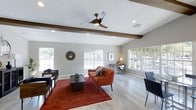 This single-floor clubhouse offers a thoughtfully designed communal space that blends comfort with modern style. The main lounge area serves as the heart of the clubhouse, featuring light hardwood-style flooring, neutral gray walls, and a crisp white ceiling accented by rich exposed wood beams that add warmth and architectural character to the room. Recessed lighting and a sleek three-blade ceiling fan with an integrated light ensure the space is well-lit and comfortable year-round. The lounge is furnished in a mid-century modern style, highlighted by a bold rust-orange sofa and matching accent chair, a dark wood coffee table set atop a vibrant red area rug, and additional seating along the perimeter. Multiple large windows line the walls, filling the interior with generous natural light and providing views of the surrounding outdoor environment. A dedicated workspace near one of the windows includes a wooden desk and ergonomic chair, making the clubhouse functional for both social gatherings and individual productivity. The overall ambiance is inviting and well-appointed, striking a balance between a relaxed residential feel and a polished community gathering space.