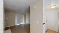 This single-story unit offers a straightforward, functional layout finished consistently with neutral warm-toned walls and tile flooring throughout. The living area is generously proportioned, featuring a sliding glass door dressed with vertical blinds that allows natural light to fill the space. A textured ceiling adds subtle visual interest overhead.

Adjacent to the living area is a dining space with a window fitted with horizontal blinds, providing additional natural light. This area connects directly to the galley-style kitchen, which is efficiently arranged with white cabinetry above and below the countertops, a coil-burner electric range, a full-size refrigerator, and a dishwasher — all in white, maintaining a clean, cohesive look.

The bedroom is a comfortable, well-lit room with a window and a dedicated walk-in closet equipped with dual hanging rods, providing ample storage. The bathroom features a tub-shower combination with a curtain rod. A notable feature of the unit is a small entry or utility area distinguished by a painted white brick accent wall, adding a distinctive architectural character to the otherwise neutral interior. The space also includes a fire extinguisher and smoke detectors, reflecting attention to safety throughout.