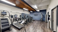 Located at 80–90 Park Street in Springfield, MA, this fitness center offers a thoughtfully organized exercise space across a single floor. The facility is divided into distinct zones, seamlessly connected through glass-paneled interior windows that allow visibility between areas while maintaining separation.

The main cardio area is a standout, featuring a row of modern treadmills, ellipticals, stair climbers, and a recumbent bike, all positioned along a full-length wall mirror that amplifies the sense of space. Exposed brick walls and warm wood-paneled ceiling beams lend a distinctive industrial-meets-modern aesthetic, while overhead fluorescent lighting ensures the area is well-lit throughout.

A dedicated strength training zone is equipped with a cable machine, adjustable weight bench, free weights, and a trampoline for dynamic training. The blue accent walls, one of which displays a bold motivational "WORKOUT" graphic, contribute to an energizing ambiance. Durable wood-look flooring runs throughout, providing both a practical and polished surface.

The space also includes a secondary room visible through glass partitions, housing additional strength equipment and a dumbbell rack. High ceilings with exposed structural beams add to the open, airy feel of the facility, making it a functional and motivating environment for a full range of fitness activities.