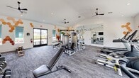 Located at 1931 Chemin Metairie Pkwy in Youngsville, Louisiana, this fitness center offers a comprehensively outfitted exercise space designed for a well-rounded workout experience. The room features vaulted ceilings that lend an open, airy feel, complemented by multiple ceiling fans that promote air circulation throughout. Crisp white walls are accented with distinctive honeycomb-patterned wall art in warm orange and brown tones, giving the space a modern, energetic aesthetic. Large windows and glass-panel doors allow natural light to pour in while providing views of the surrounding neighborhood landscape. The flooring combines plush carpet tiles in a gray toned pattern with rubber mat sections designated for functional training. Equipment is varied and well-distributed, including a multi-station cable and weight machine, adjustable benches, a full dumbbell rack with wall-mounted flat-screen TV above, a power rack loaded with accessories, rowing machines, and a stationary bike. A large mirror along one wall amplifies the sense of space and supports form-checking during workouts. Built-in shelving provides convenient storage, and restroom access is available on-site. The facility is bright, functional, and thoughtfully designed to support a broad range of fitness activities.