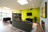 Located at 3955 Marina Lake Road in Virginia Beach, Virginia, this single-story community clubhouse offers a vibrant and welcoming gathering space designed for both relaxation and socializing. The open-plan layout is anchored by multiple distinct lounge areas, each thoughtfully furnished and styled with bold accent colors, including a striking chartreuse feature wall and soft lavender and gray tones that give the space a modern, energetic character.

Light wood-look flooring flows throughout, unifying the various seating zones beneath recessed lighting and skylights that keep the interior bright and airy. A standout feature is the expansive floor-to-ceiling window wall that frames sweeping waterfront views of the adjacent lake, flooding the main lounge with natural light and providing a serene backdrop. French doors along the perimeter further connect the interior to the outdoors.

The main seating area is generously sized, featuring plush sectional and accent seating arrangements ideal for gathering. A secondary lounge area with a wall-mounted television and cozy sofa configuration offers a more intimate setting. The space also includes a well-equipped kitchen with ample counter space, warm wood cabinetry, dark laminate countertops, and full appliances, making it suitable for community events and entertaining. Overall, the clubhouse delivers a comfortable, stylish, and functional environment.